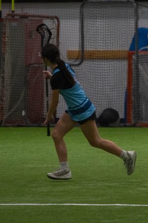 Player in blue and black uniform running with lacrosse stick during indoor practice.