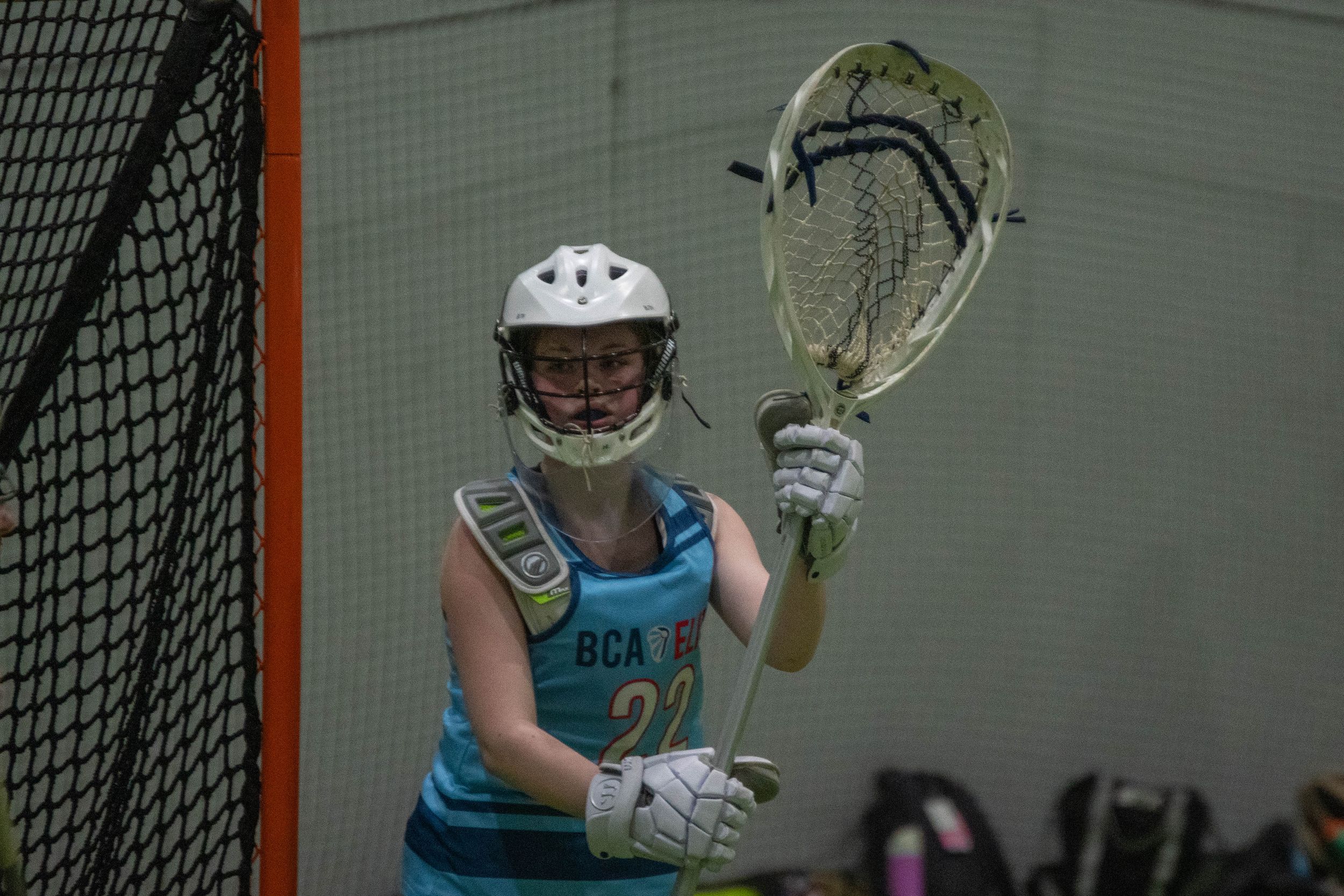 Goalie in protective gear holds lacrosse stick in front of net.