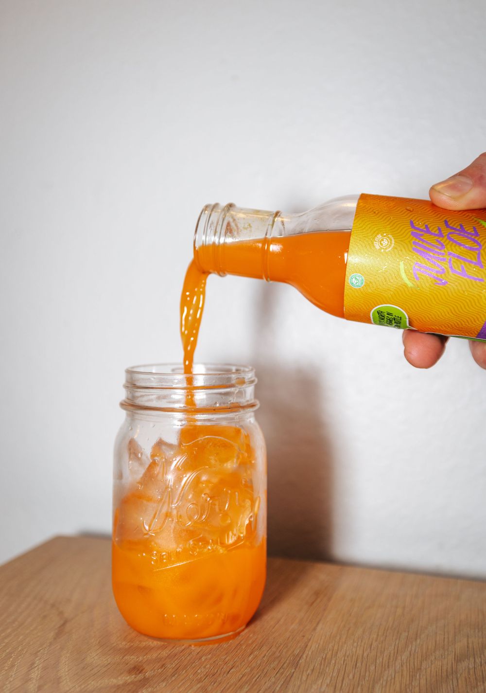 Orange beverage being poured from a bottle into a mason jar on a wooden surface.