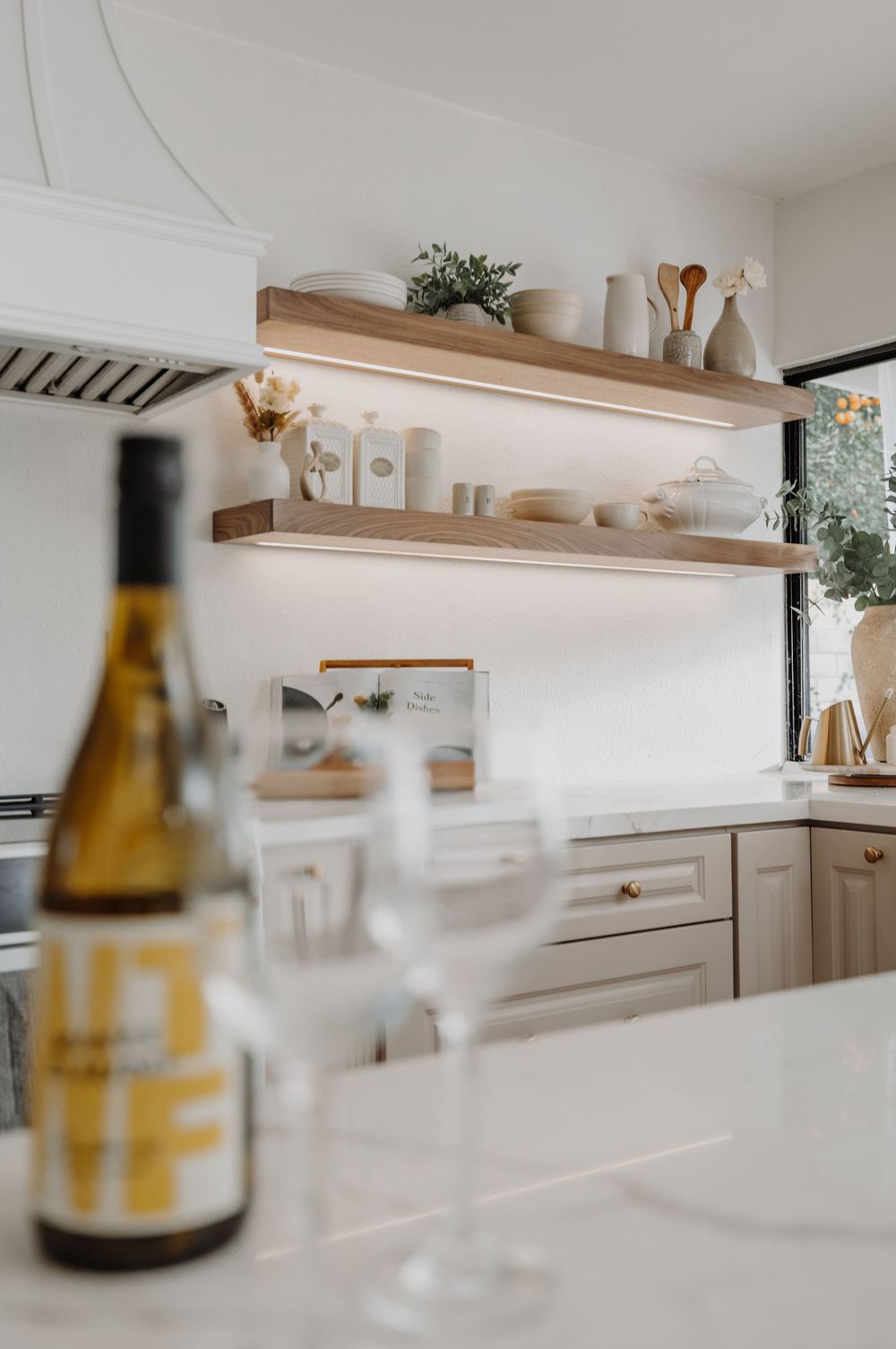 Modern white kitchen with floating wooden shelves and decorative glassware.