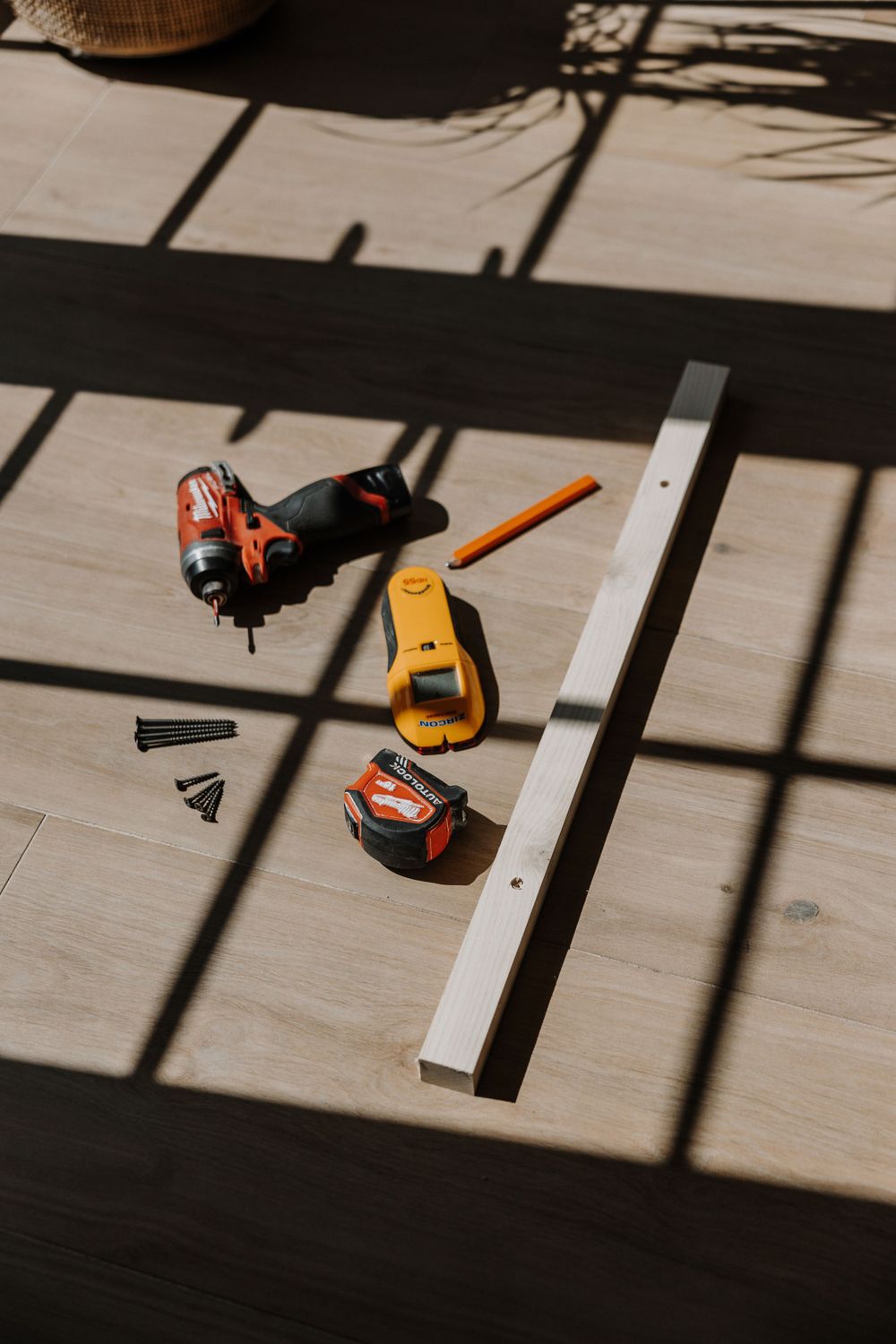 Woodworking tools and measuring tape scattered on a light wooden surface with dramatic window shadows.