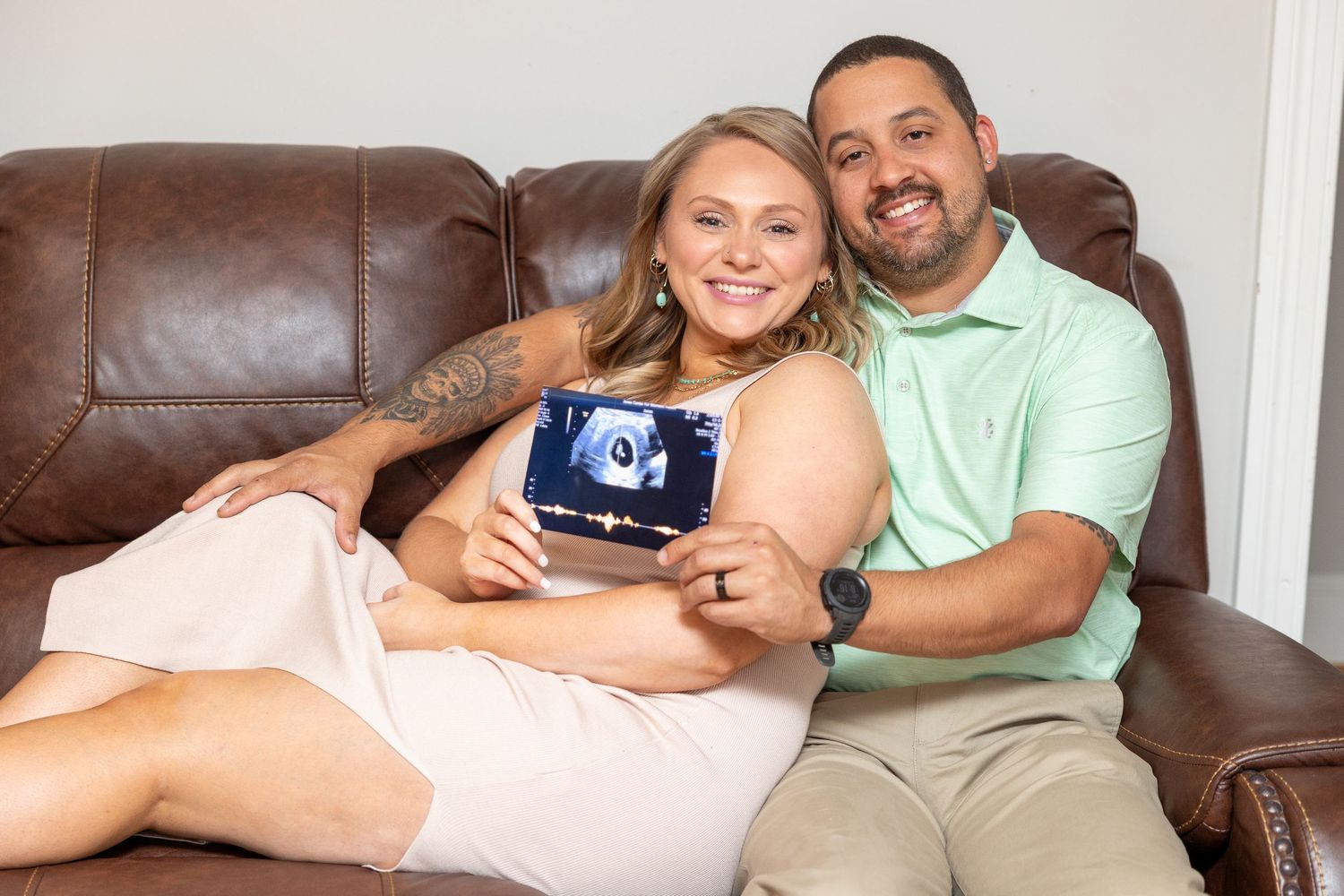 Couple relaxing together on brown leather couch at home while showcasing their ultrasound of new baby (Clinton, NC).
