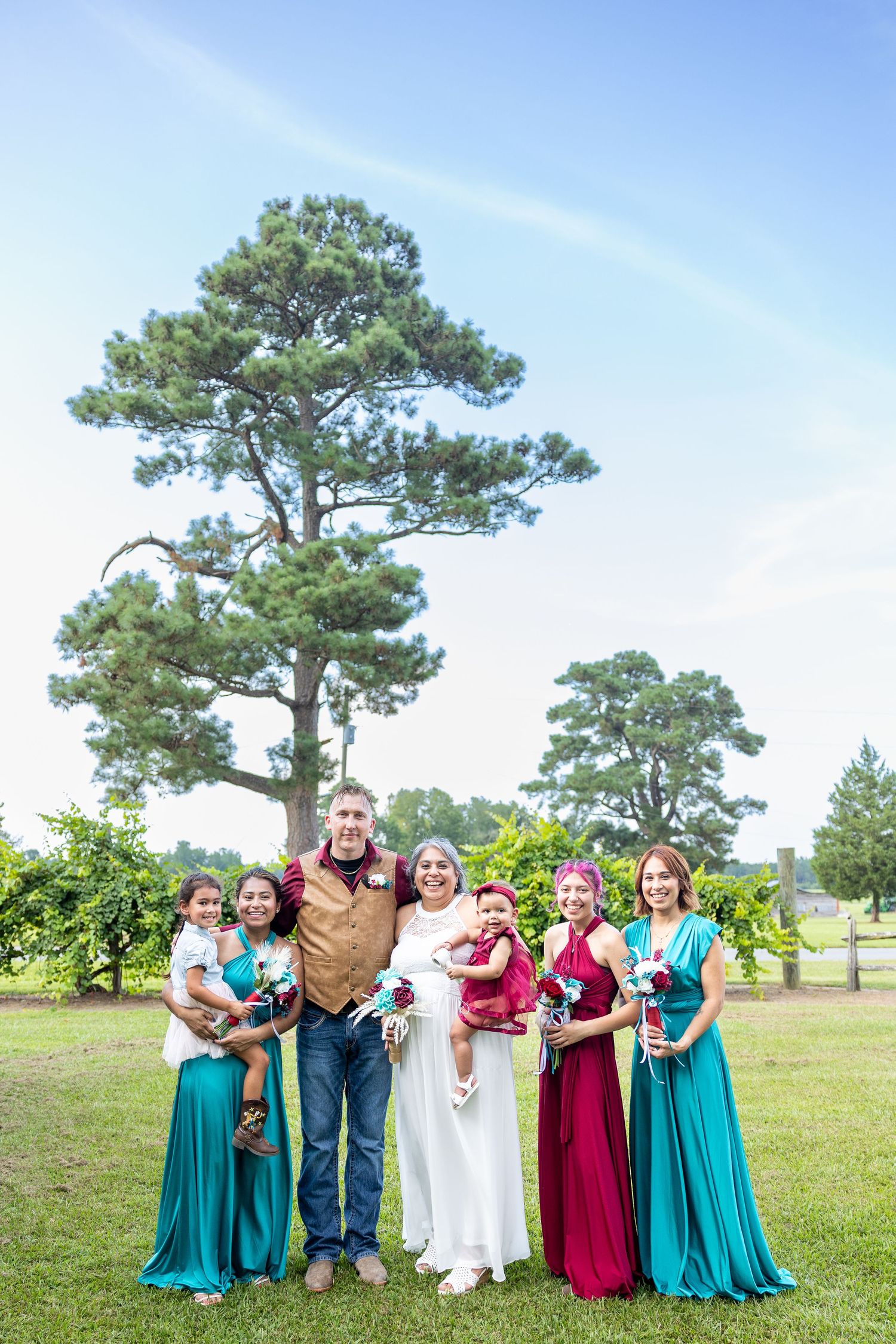 Wedding party poses together outdoors under tall pine trees wearing teal and burgundy dresses (Beulaville, NC).