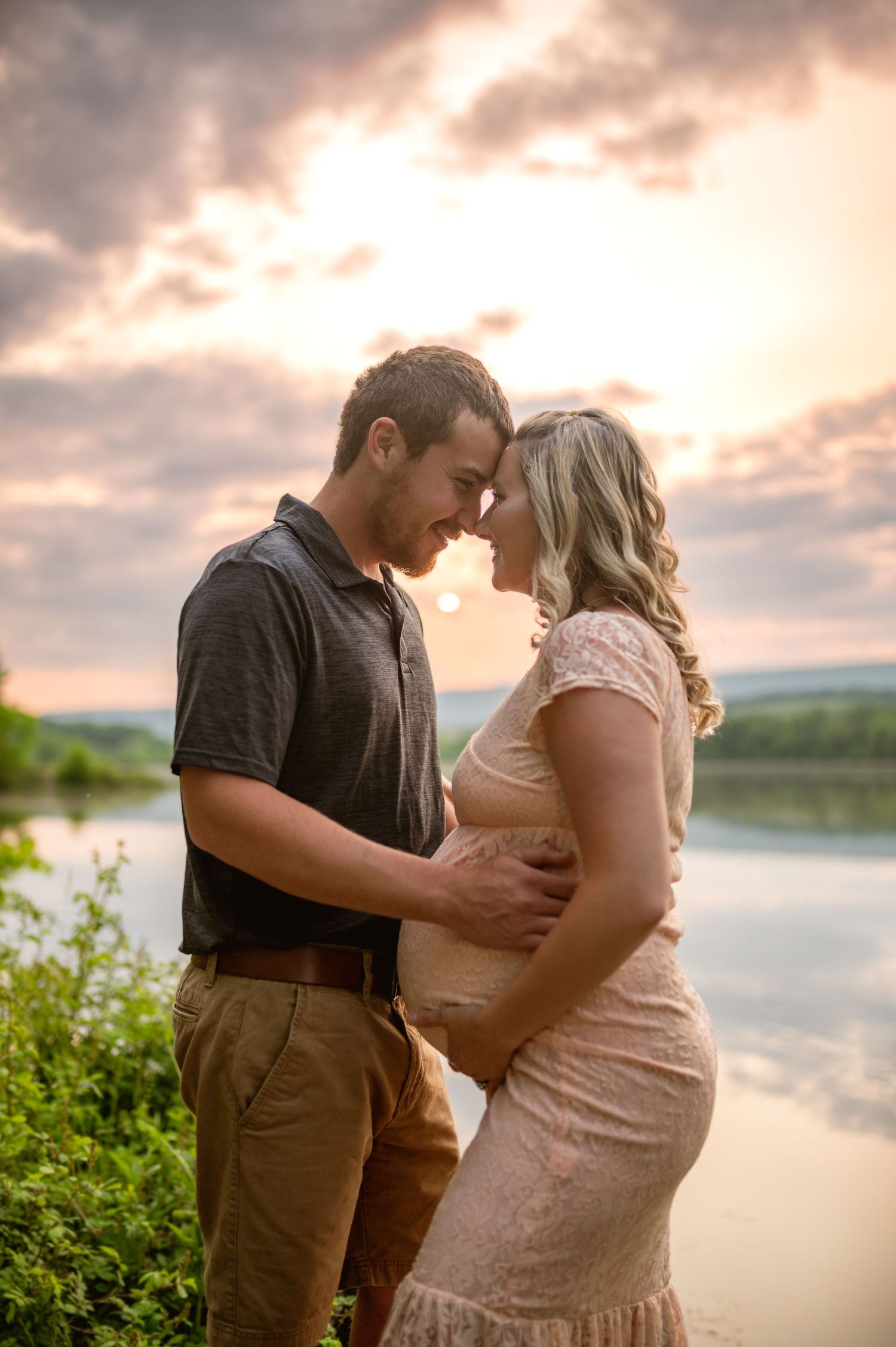 Romantic couple sharing an intimate moment at sunset by a tranquil lake, with dramatic clouds in the background.