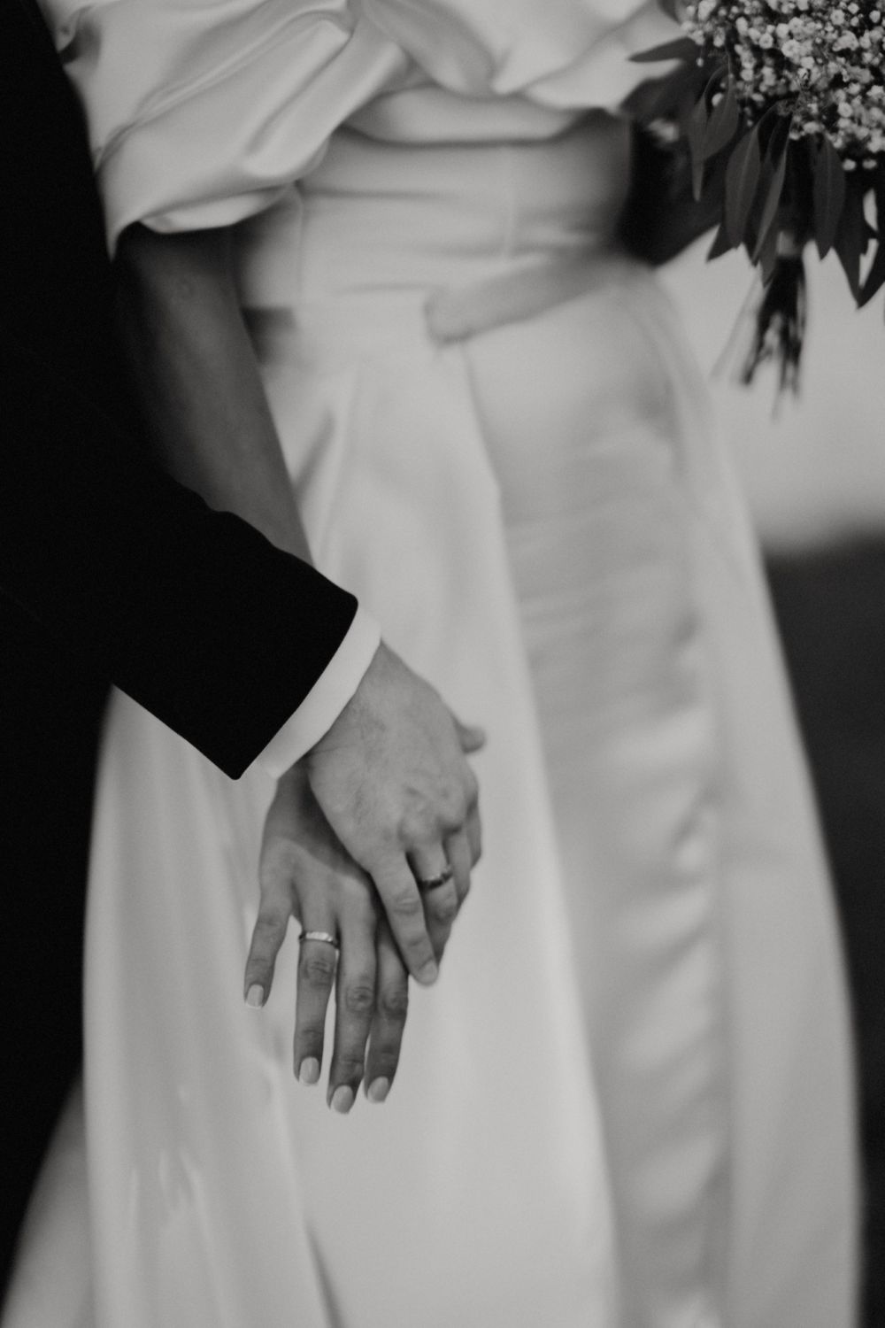 Intimate black and white detail shot of wedding rings and joined hands against white dress.