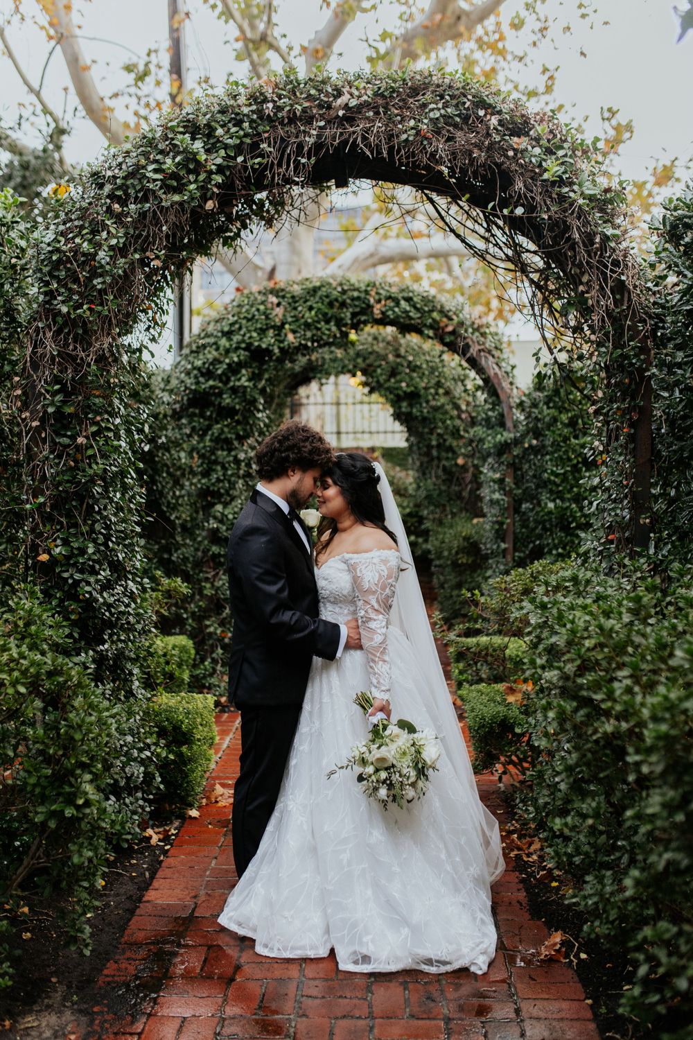 Wedding couple sharing intimate moment under lush garden archway with brick pathway.