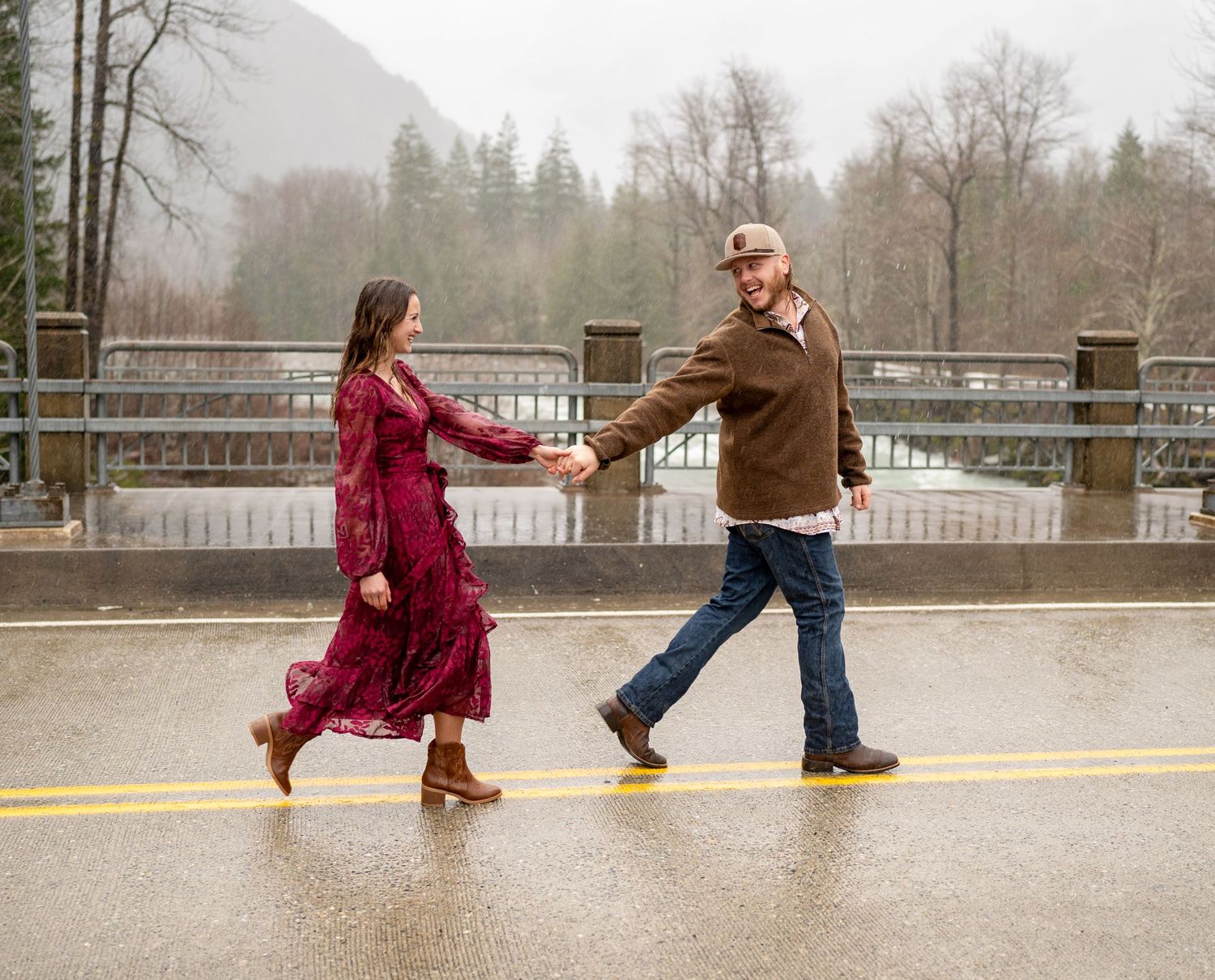 Two people dance playfully in the rain on a misty mountain road with a bridge railing and evergreen trees in background.