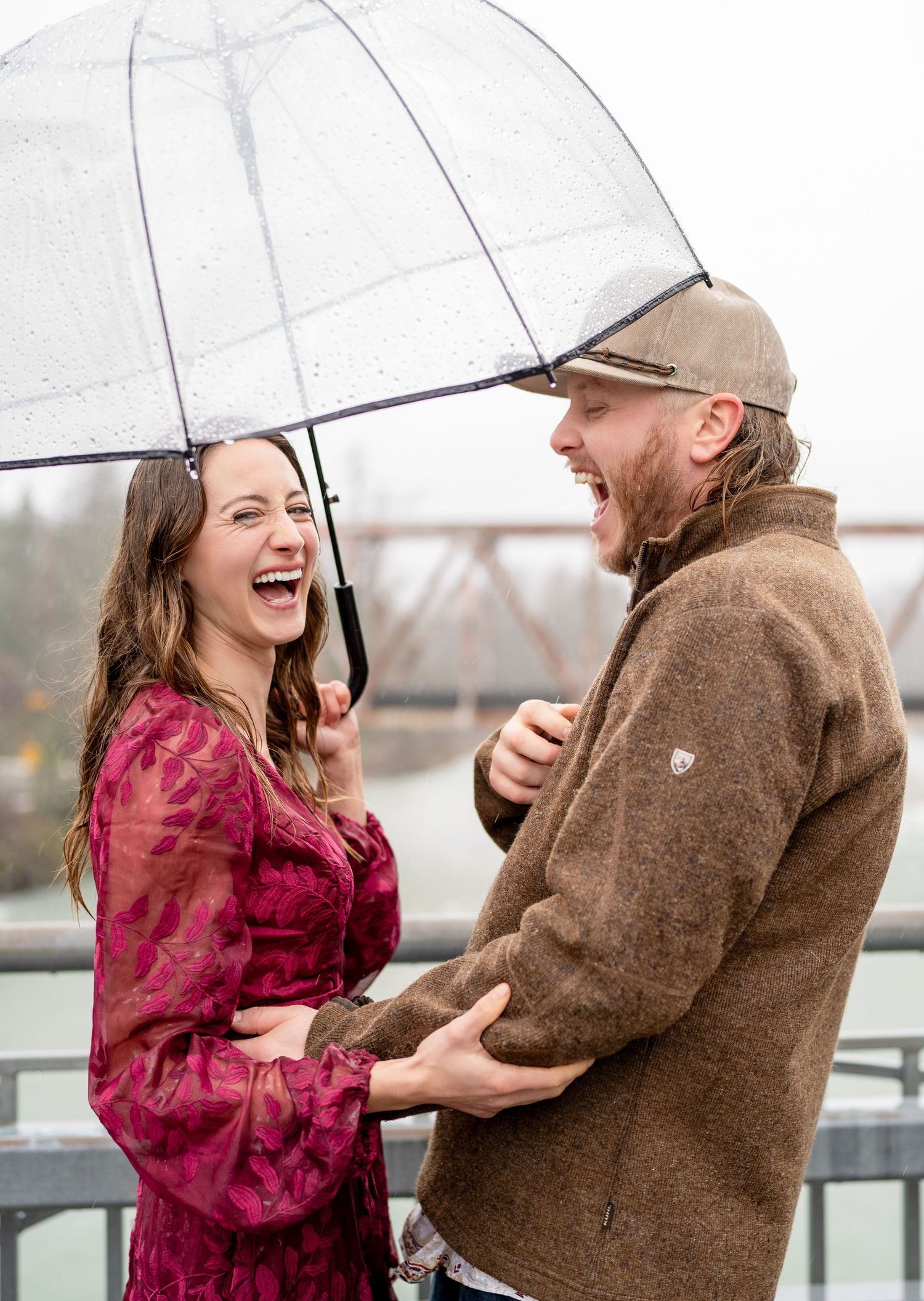 A couple shares a joyful laugh together under a clear umbrella on a rainy day by a bridge.