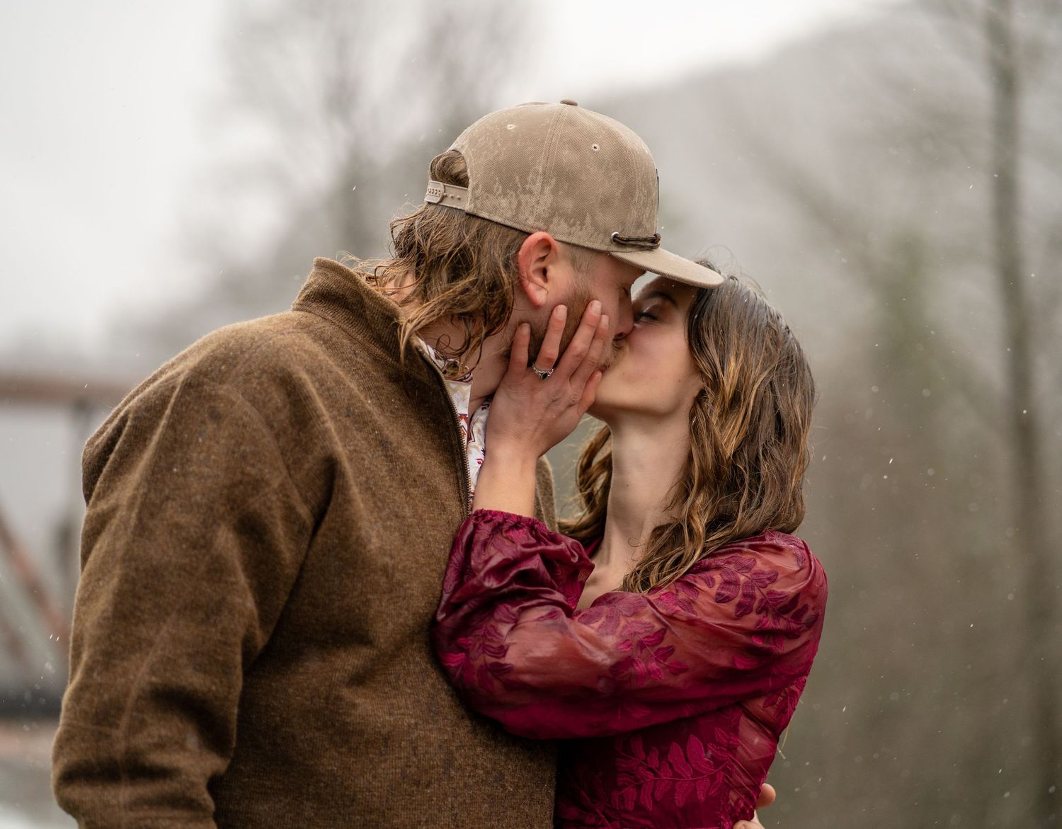 A couple embraces in a romantic kiss outdoors on a misty winter day, wearing a brown coat and burgundy sweater.