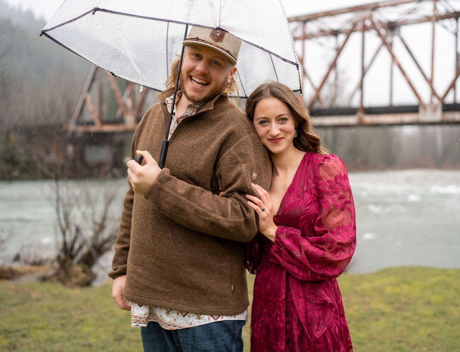 A couple poses together with an umbrella on a rainy day by a steel bridge over a river in winter.