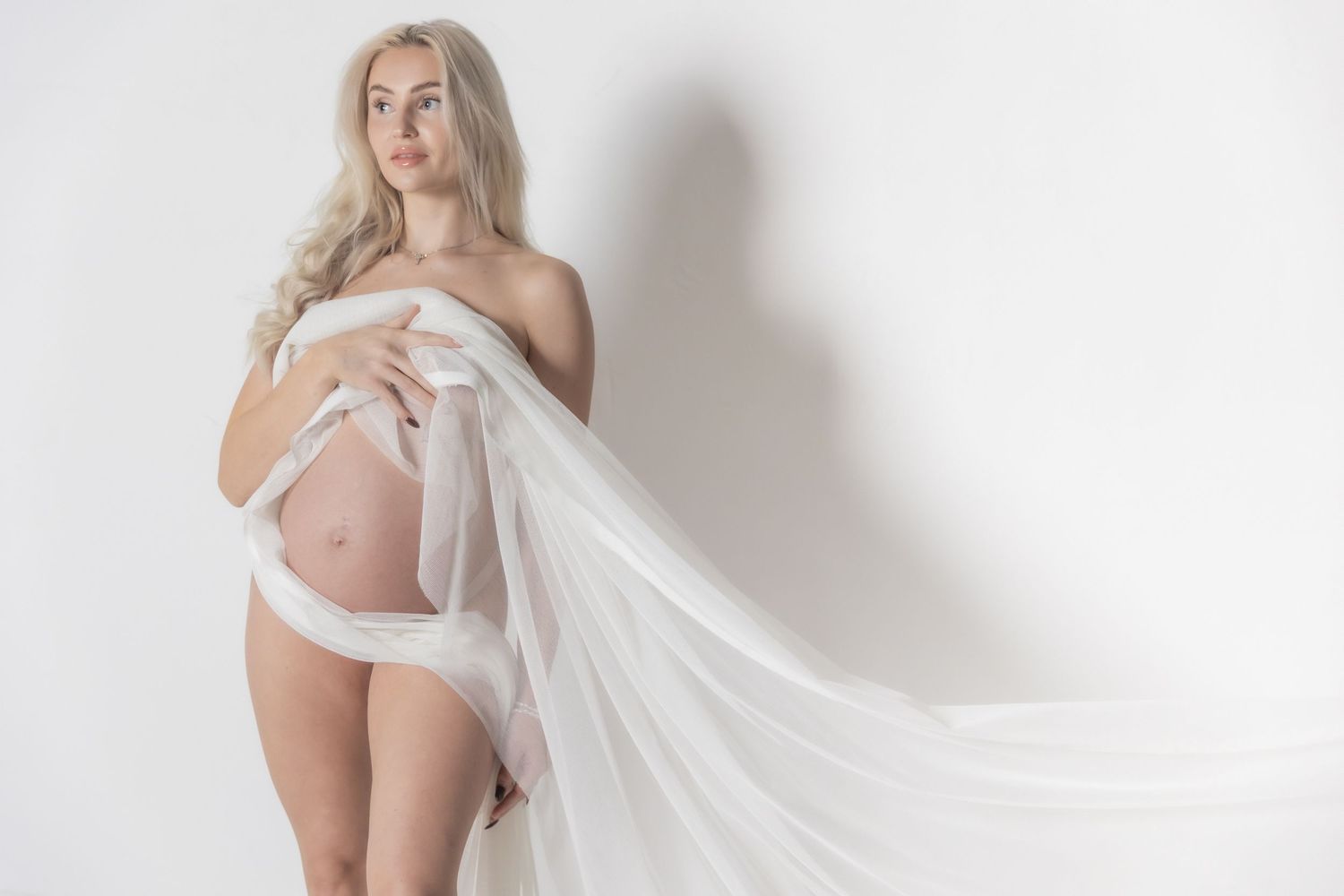 Pregnant woman posing elegantly with flowing white fabric draped around her baby bump in a studio.
