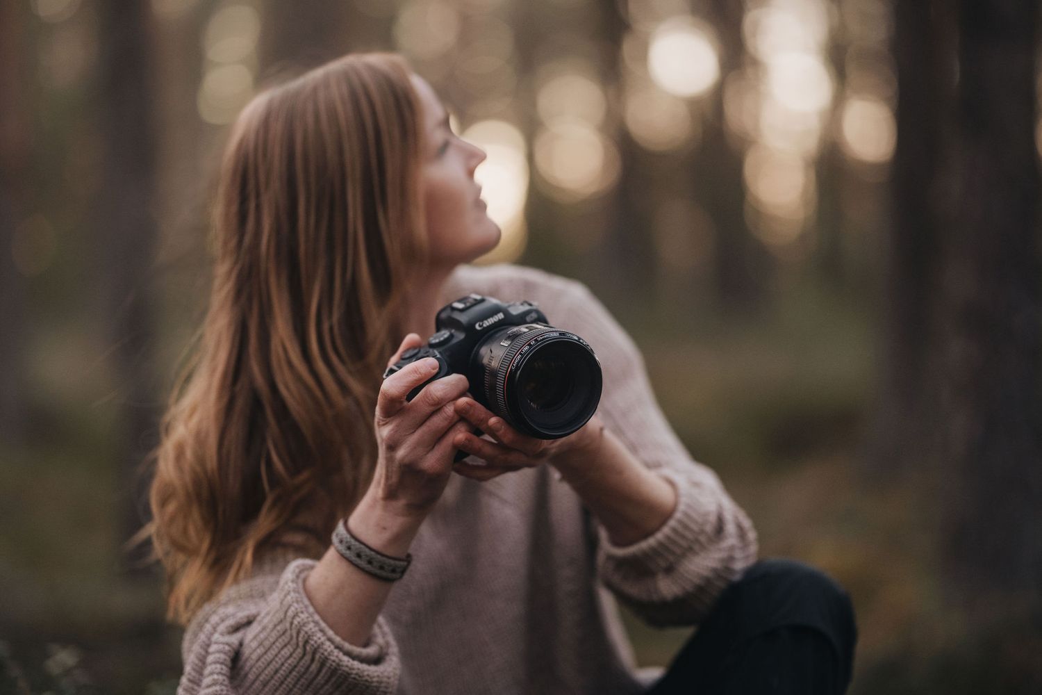 Young woman holding DSLR camera looking up in bokeh forest light.