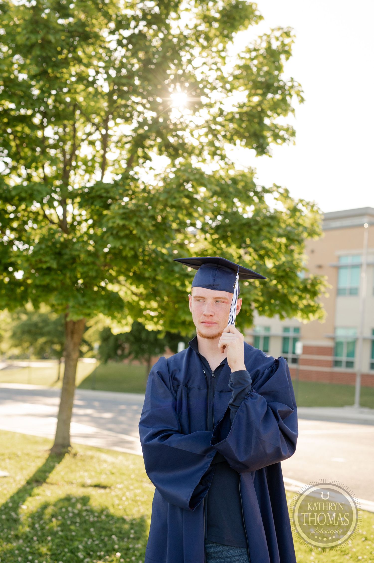 Graduate in cap and gown poses thoughtfully in golden evening light with trees behind.