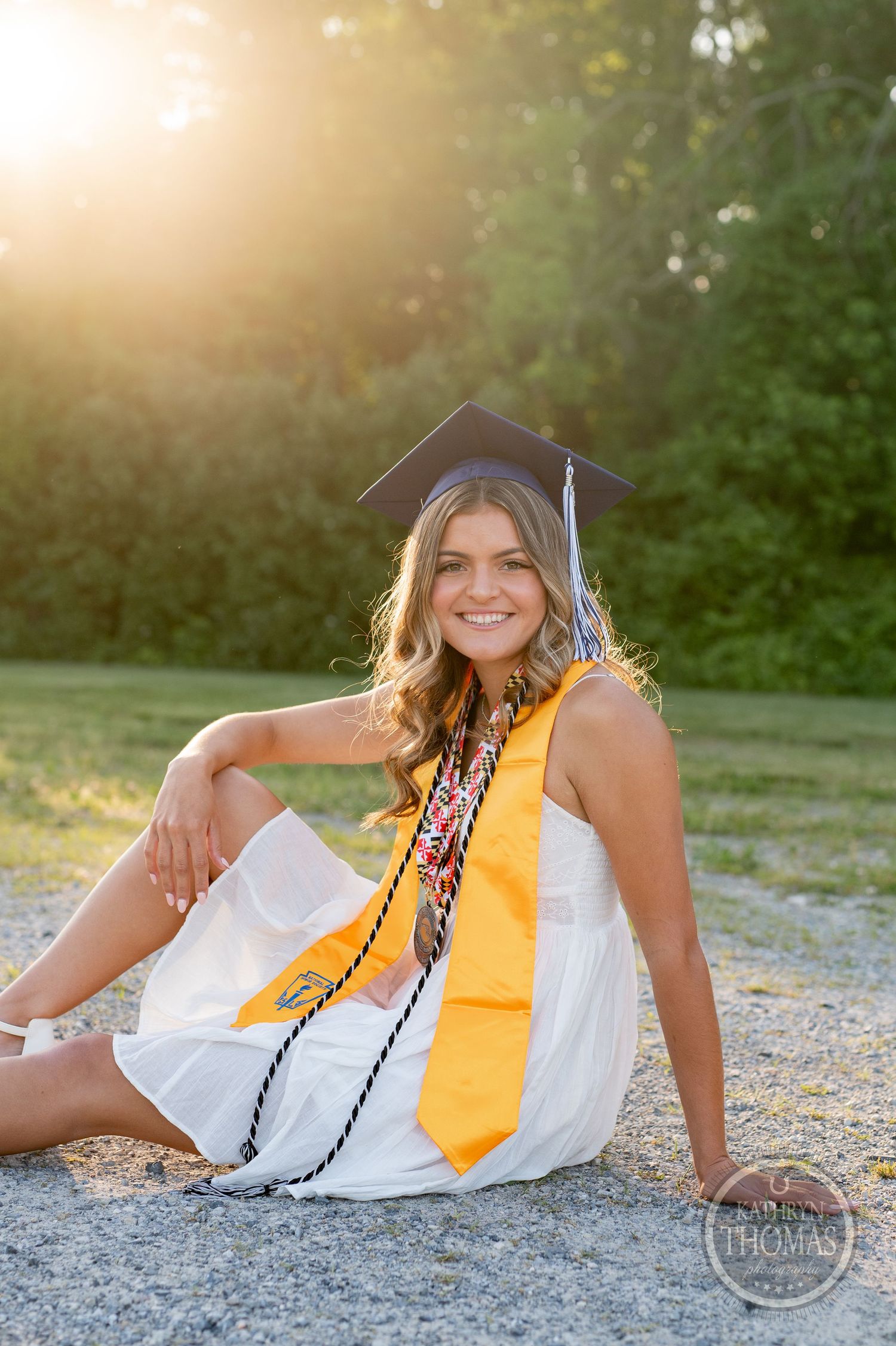Graduate in white dress with yellow stole sits on ground during golden hour photoshoot.