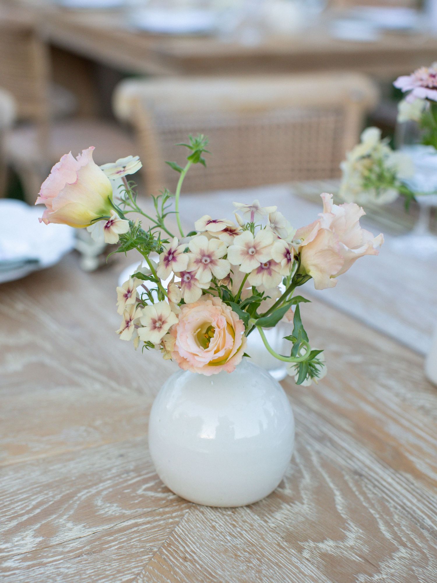 A row of small white porcelain vases each holding delicate peach ranunculus and wildflower arrangements.