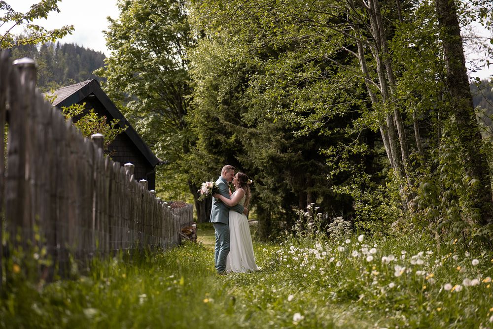 Ein Hochzeitspaar küsst sich zärtlich an einem rustikalen Holzzaun inmitten der üppigen grünen Natur des Schwarzwalds am Titisee