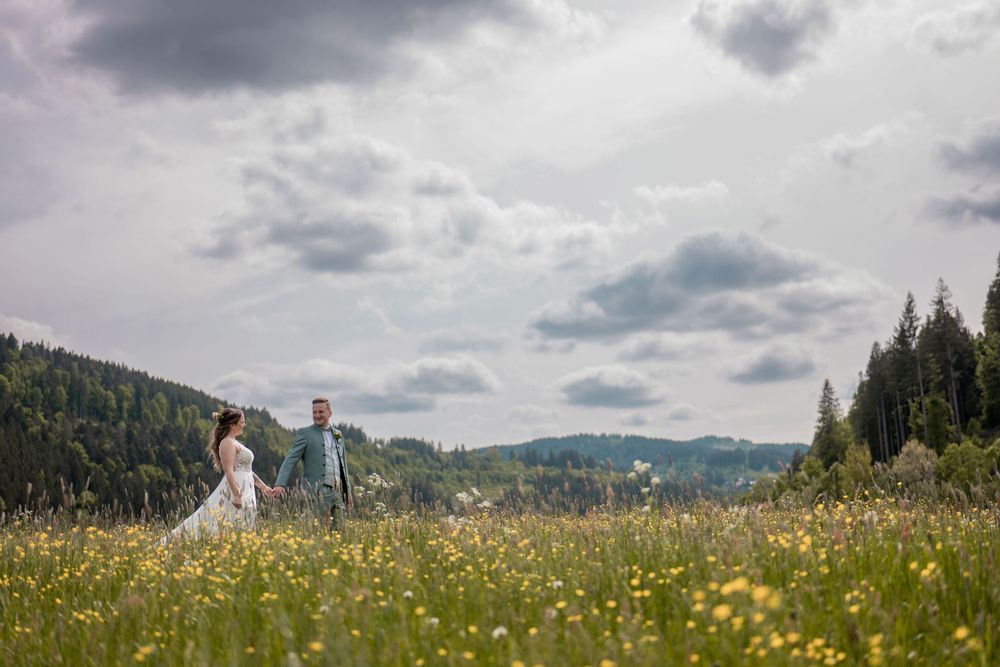 Weitwinkelaufnahme eines Brautpaares in einer weitläufigen Blumenwiese unter einem dramatischen Wolkenhimmel im Schwarzwald.
