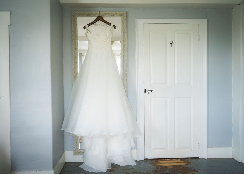 Elegant white layered wedding gown hanging on a door in a bright, softly lit room before the ceremony.
