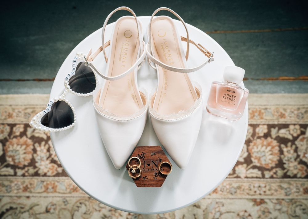 White wedding heels, heart sunglasses, gold rings in wooden box, and pink perfume bottle arranged on a white table.