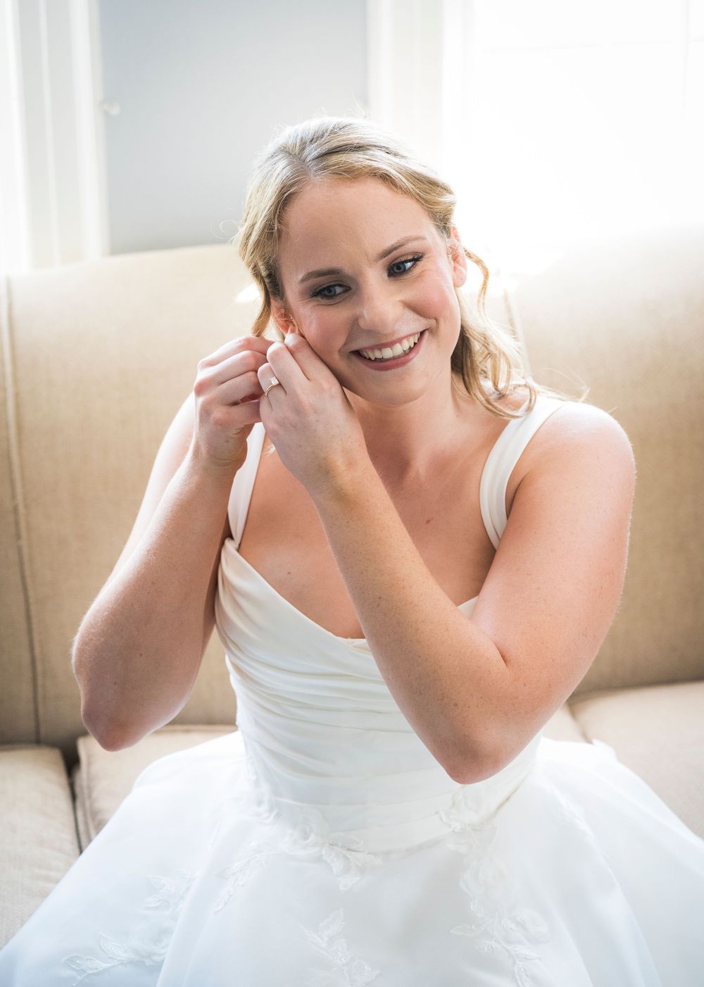 Smiling bride in white wedding gown putting on earrings while seated on a beige sofa.