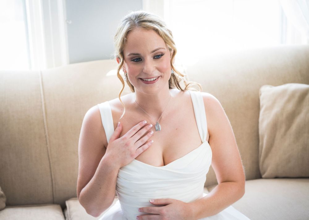 Smiling bride in white wedding dress sitting on a beige sofa touching her chest on her wedding day.