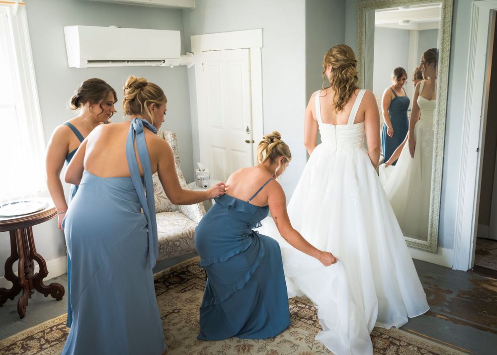 Bridesmaids in blue dresses helping the bride with her white wedding gown in a bright room.