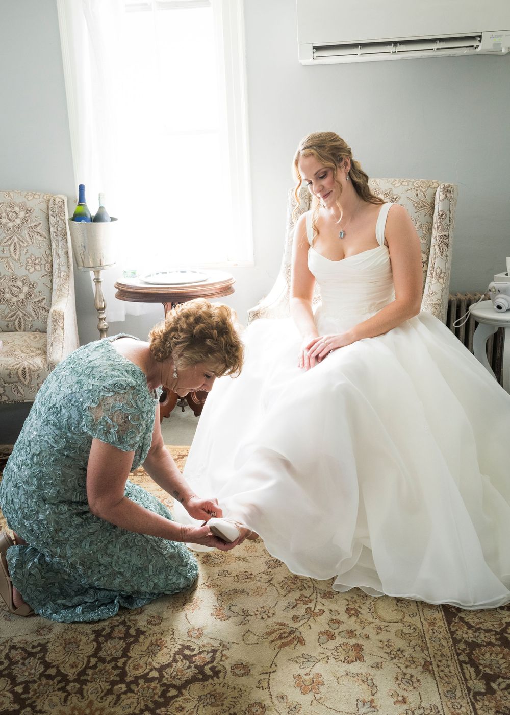 Mother helps bride with her shoes as she sits in a chair in her white ball gown before the wedding.