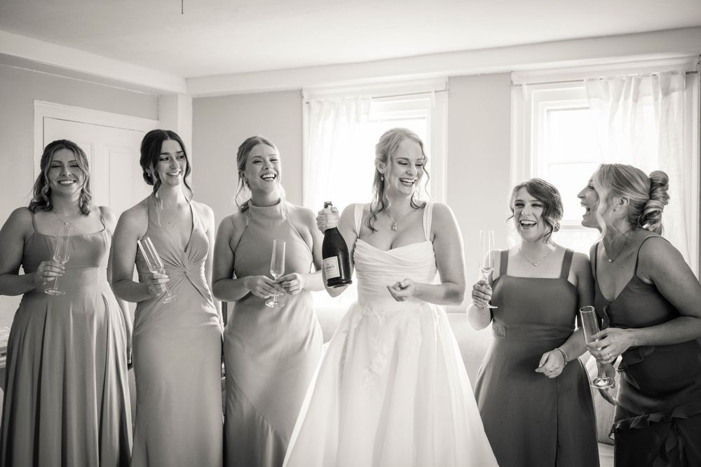 Black and white photo of smiling bride and bridesmaids in matching dresses celebrating with champagne bottles.
