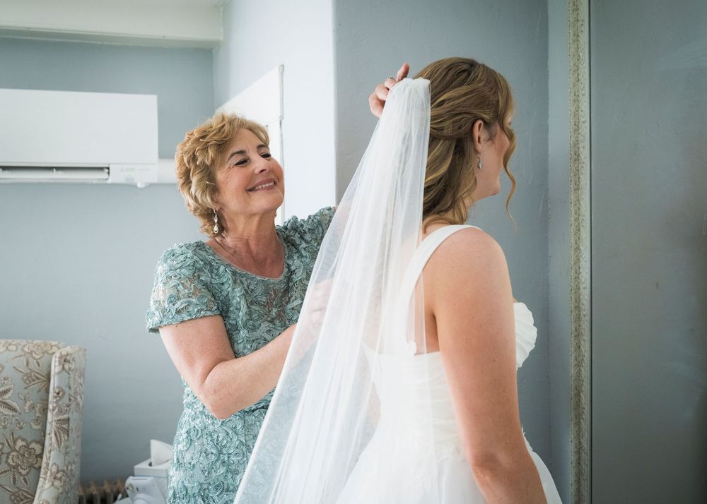 Mother helps bride adjust wedding veil during bridal preparation in bright dressing room.