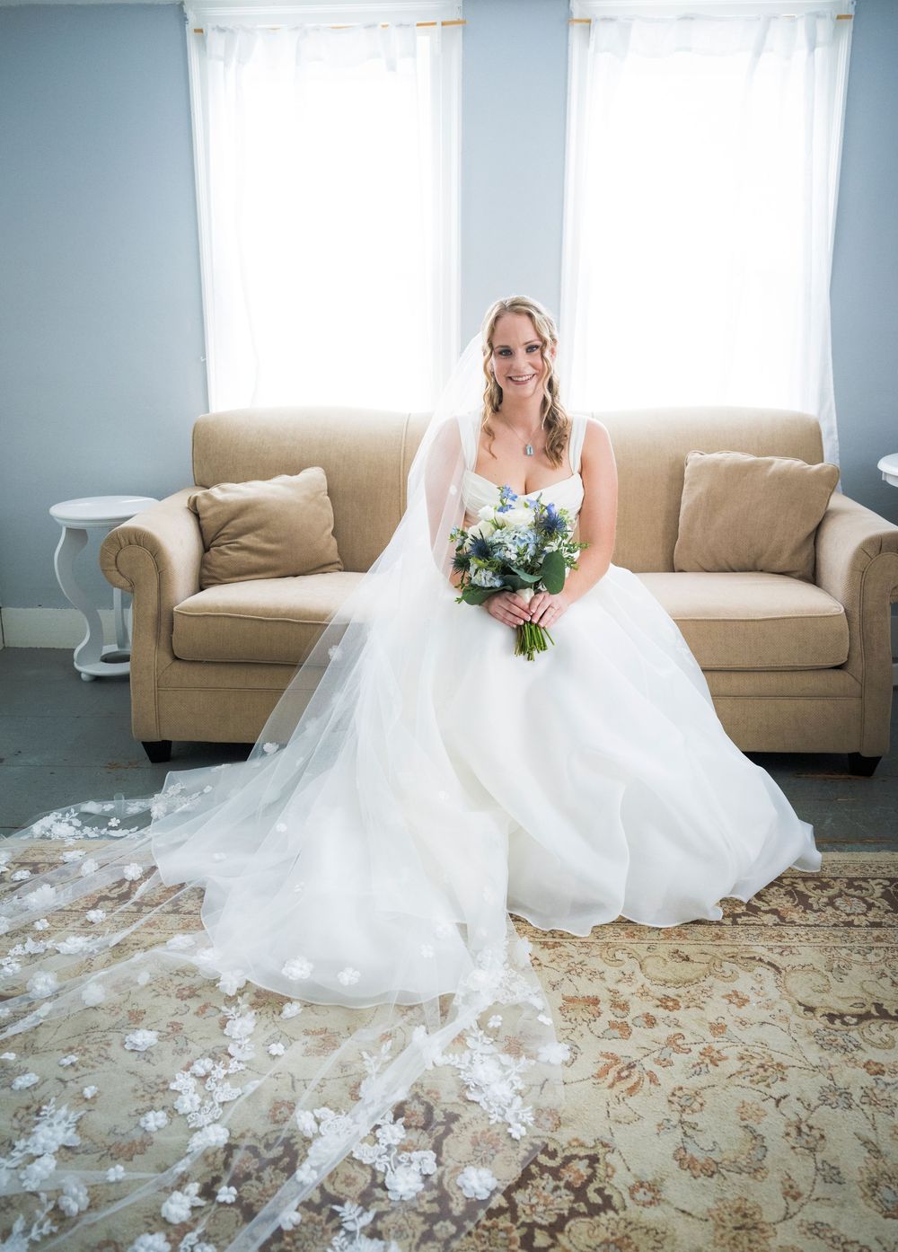Smiling bride in a ballgown with a floral veil sits on a couch holding a blue and white bouquet.