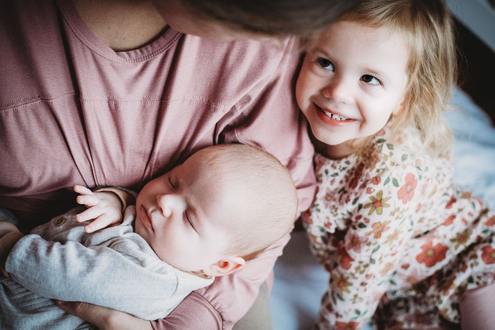 A young child in a floral dress smiles while looking at a sleeping baby resting on someone's lap.