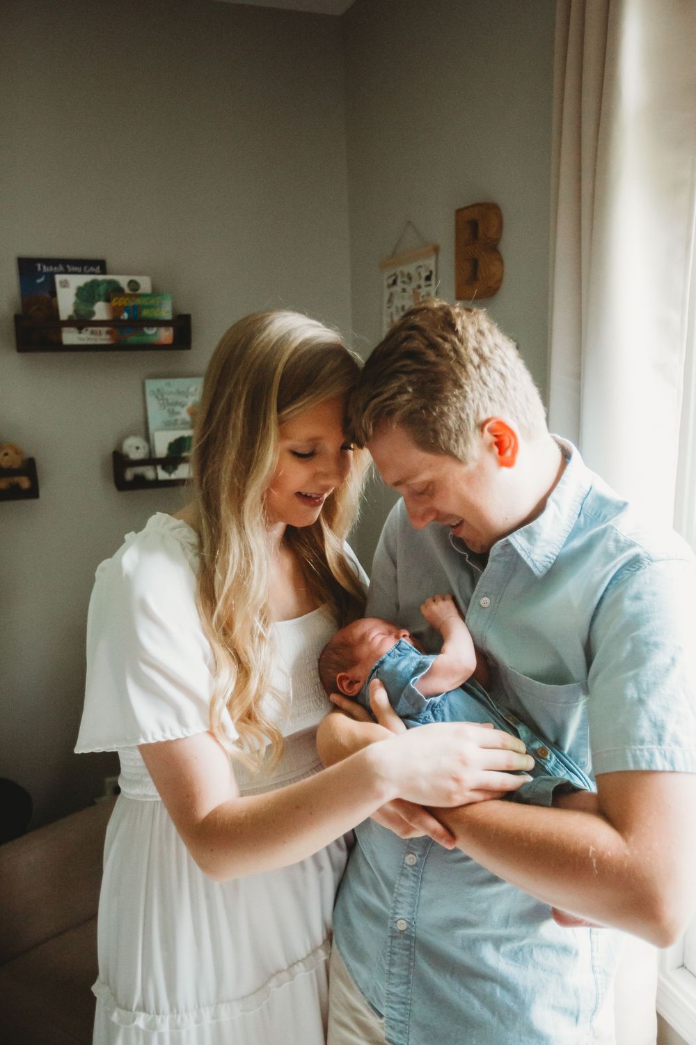 Couple in white and blue outfits sharing an intimate moment while holding their newborn baby in a nursery.