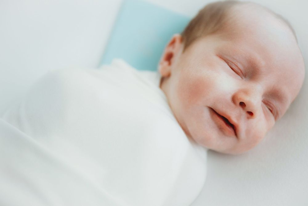A sleeping baby in white bedding smiles peacefully while napping.