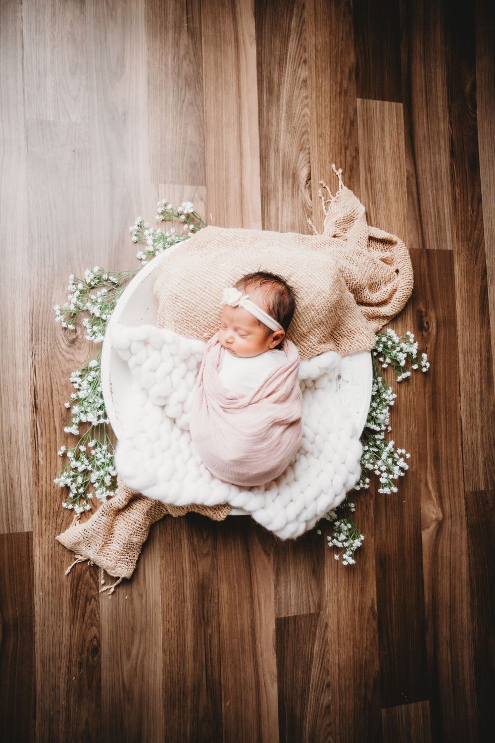 Newborn baby wrapped in soft pink swaddle lying on white blanket with delicate baby's breath flowers on wooden floor.