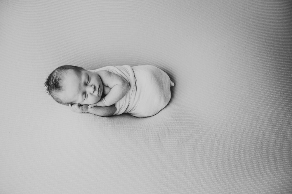 Black and white photograph of a sleeping newborn baby curled up on a soft textured blanket.
