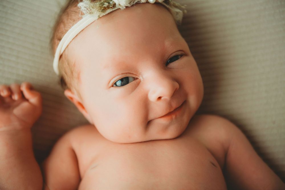 A smiling infant wearing a delicate lace headband lies on soft bedding.