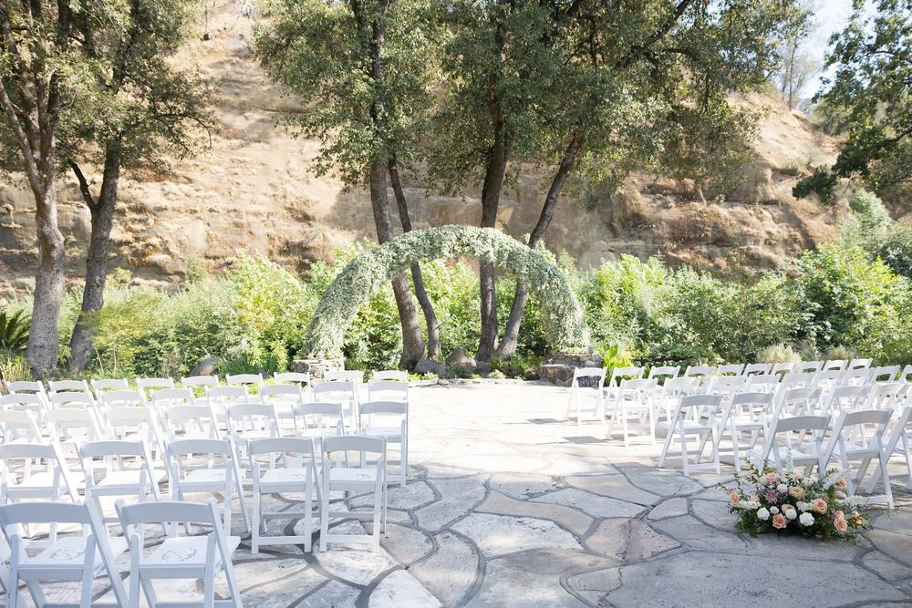 White chairs arranged for an outdoor wedding ceremony with floral arch and natural hillside backdrop.
