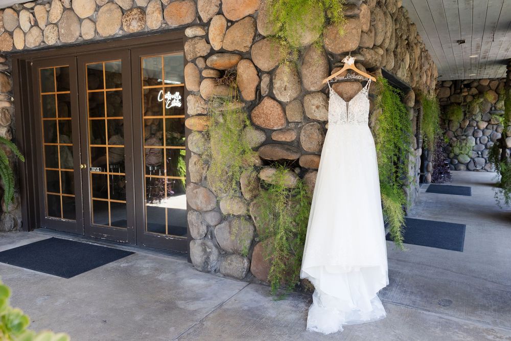 White wedding dress hangs elegantly against rustic stone wall with windows and green foliage.