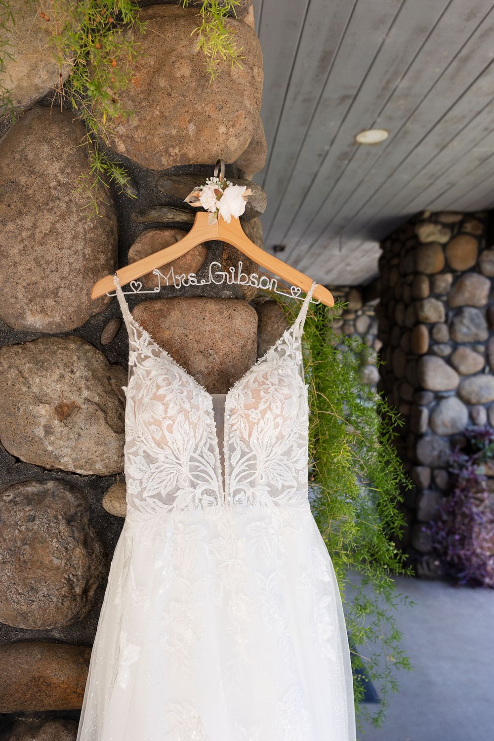 Elegant lace wedding dress with plunging neckline hangs on display against stone wall backdrop.