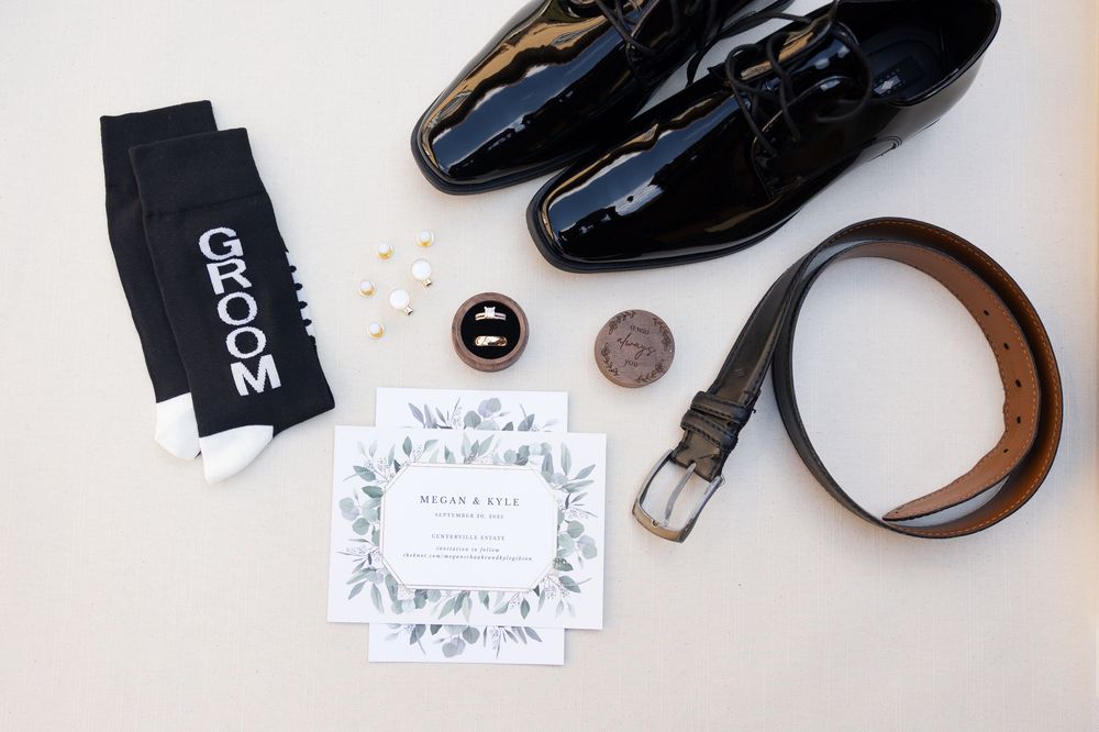 Wedding accessories including black dress shoes and paper goods arranged on white surface.