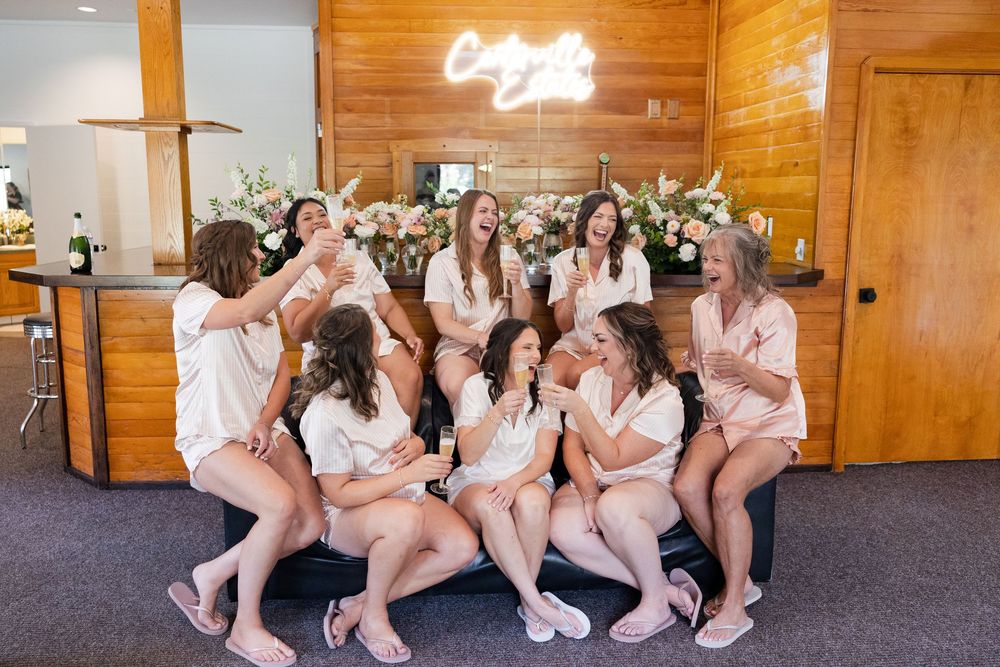 Bridesmaids in matching robes sit together on a wooden floor with neon signage in the background.