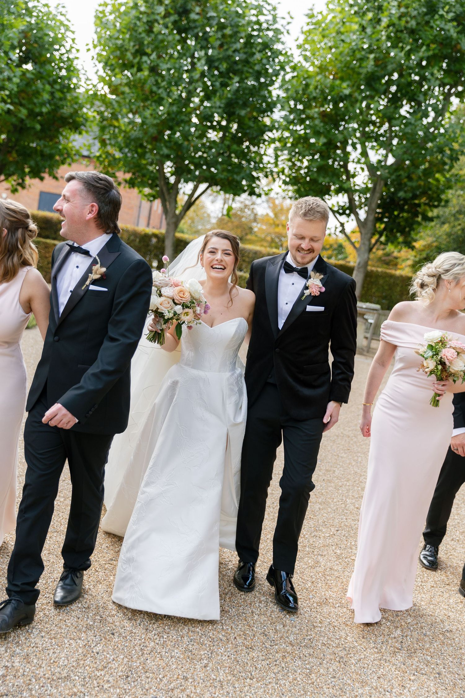 Wedding party in formal attire laughs together in candid sequence on gravel path outdoors.
