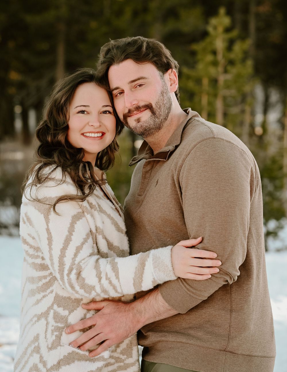 A couple embraces outdoors in winter, wearing cozy sweaters against a snowy forest backdrop.