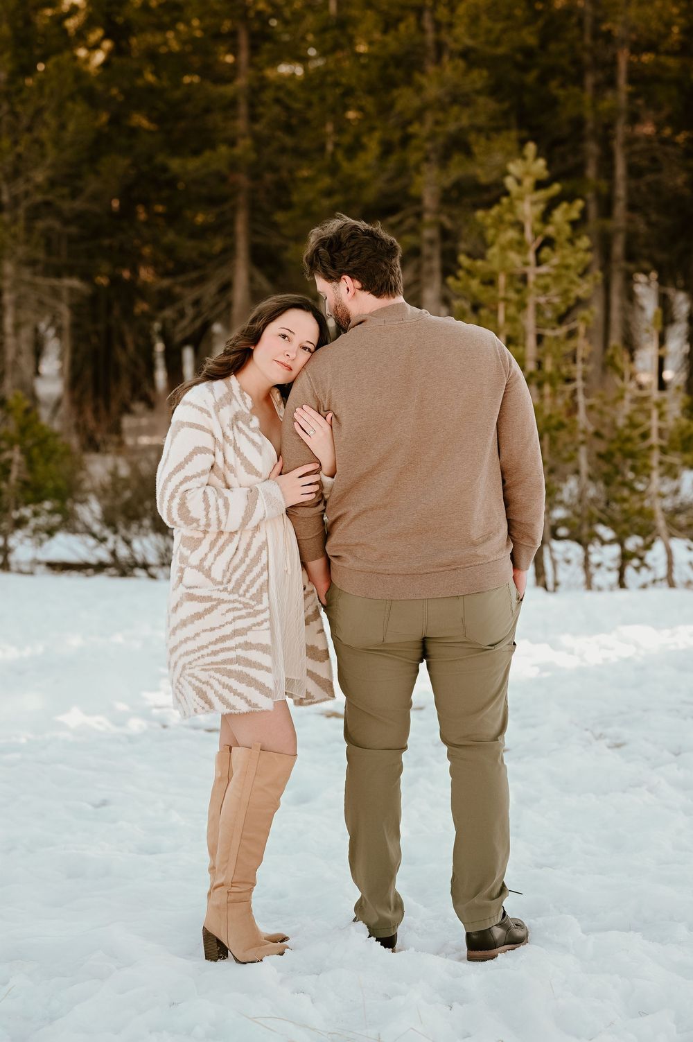 A couple stands together in a snowy winter landscape surrounded by evergreen trees in the background.