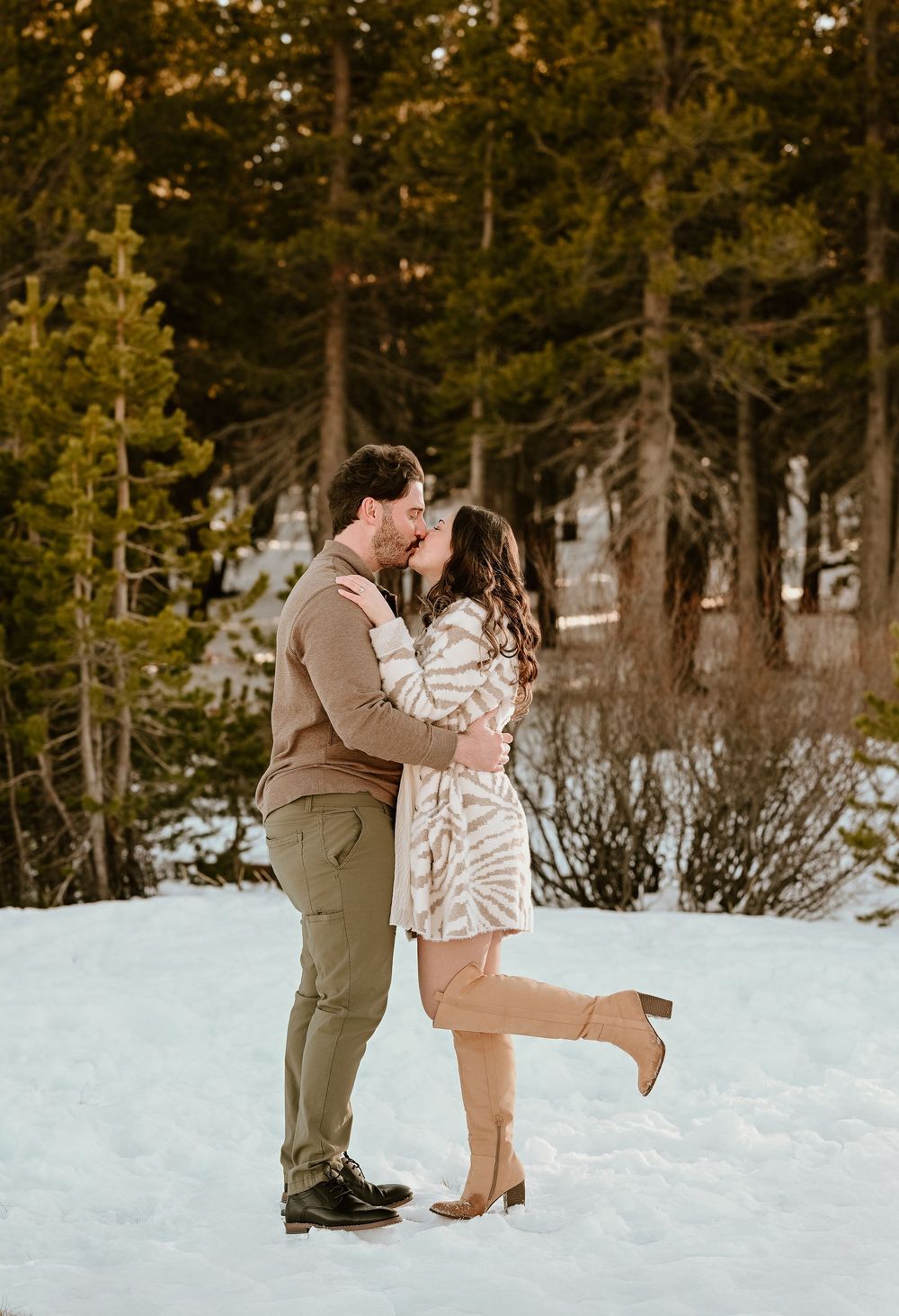 A couple shares a romantic moment in the snowy mountains with evergreen trees in the background.