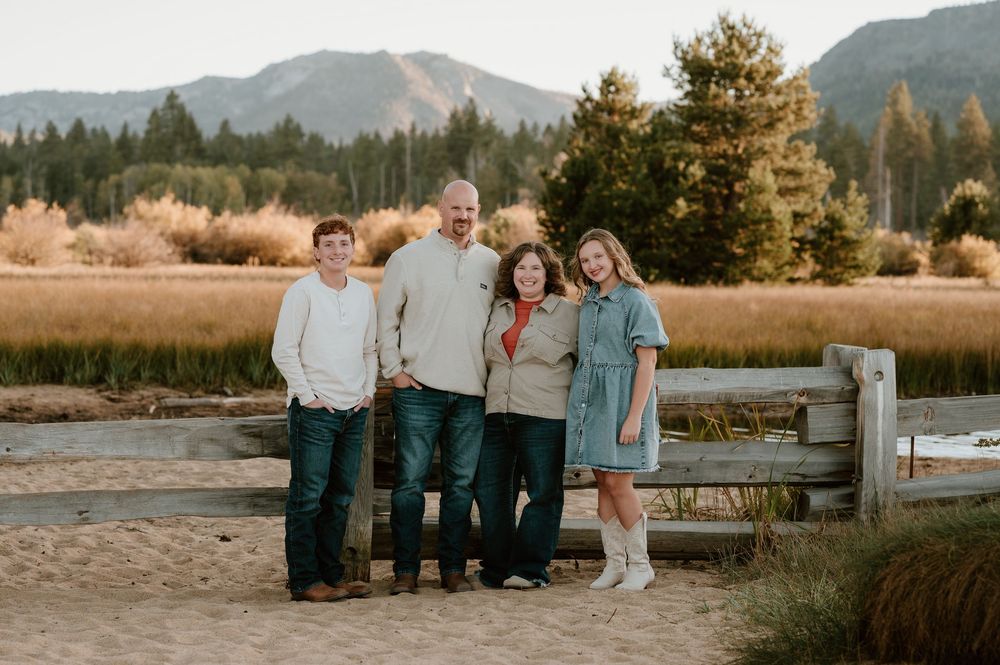 A group of people pose together at sunset near a lake with mountains and trees in the background.