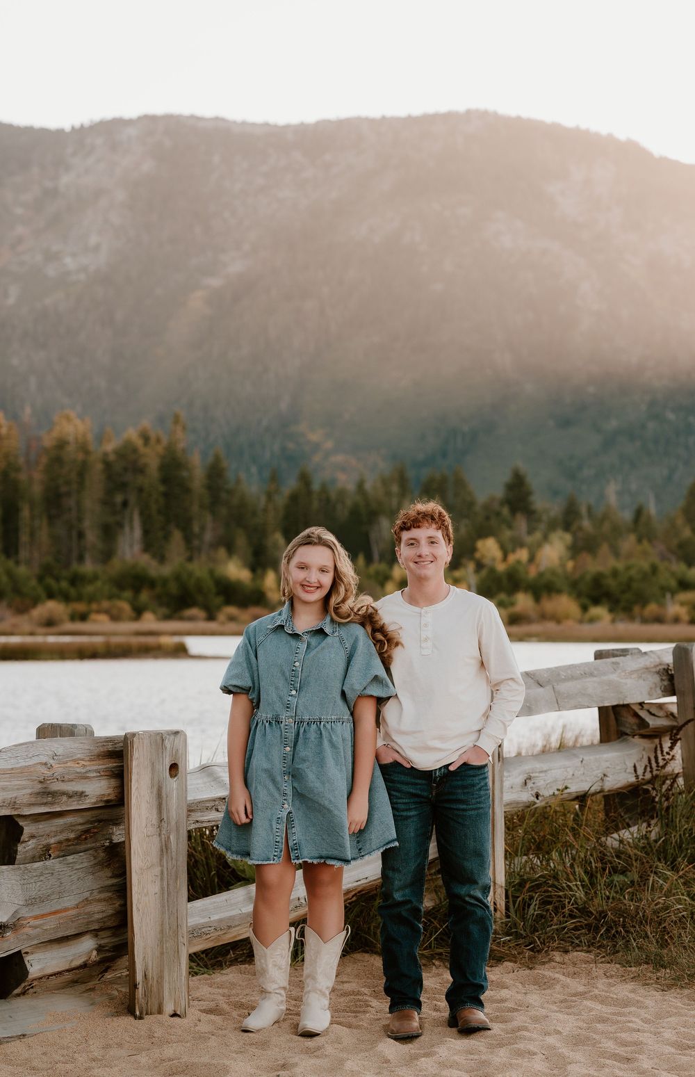 A couple stands on a wooden boardwalk near a lake with mountains and autumn trees in the background at sunset.