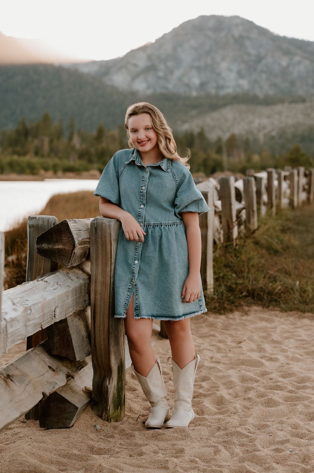 Child in denim dress and white boots sits on wooden fence at sandy beach with mountains in background.
