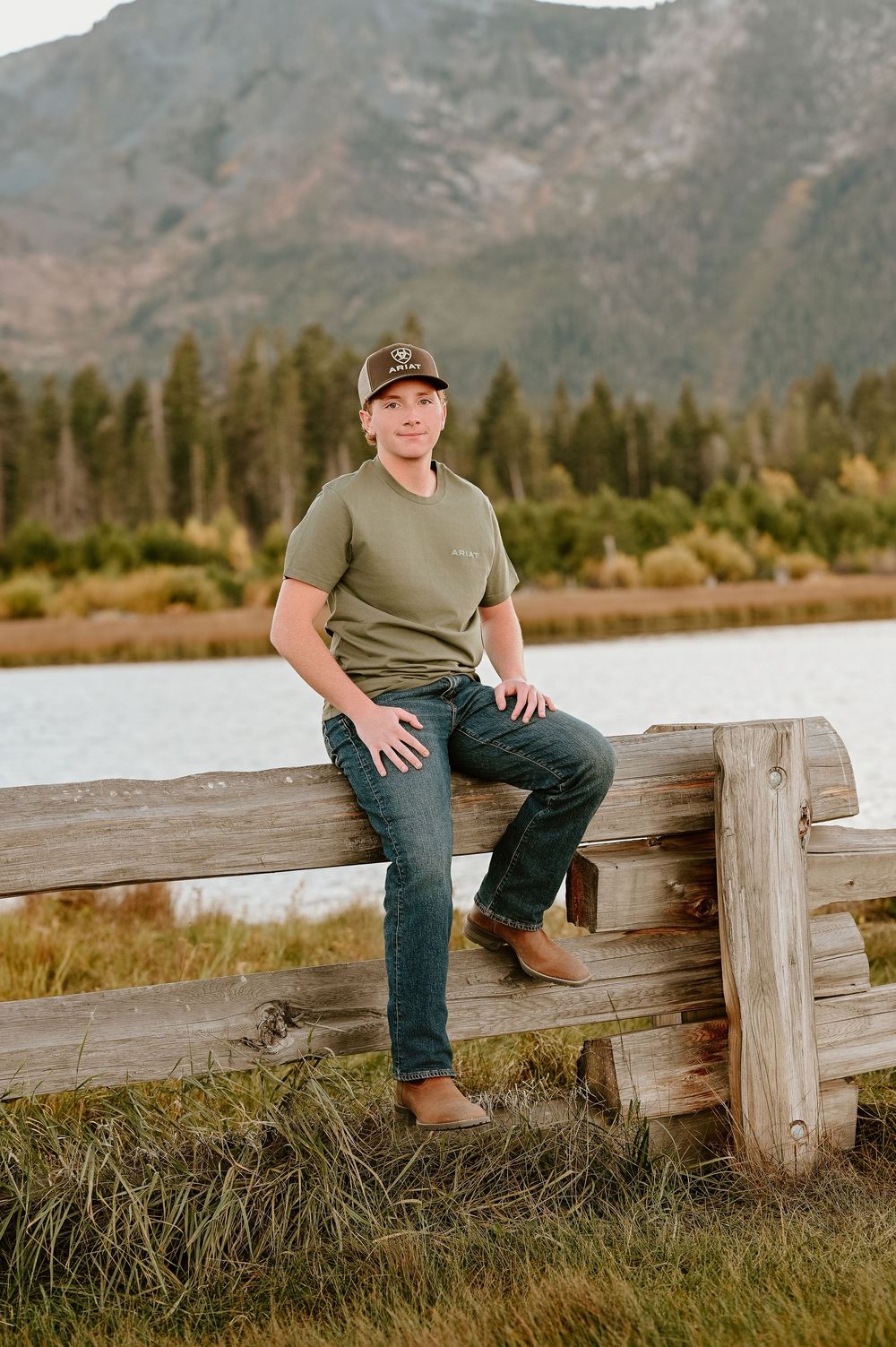 Someone sits casually on a rustic wooden fence with mountains and evergreen forest in the scenic background.