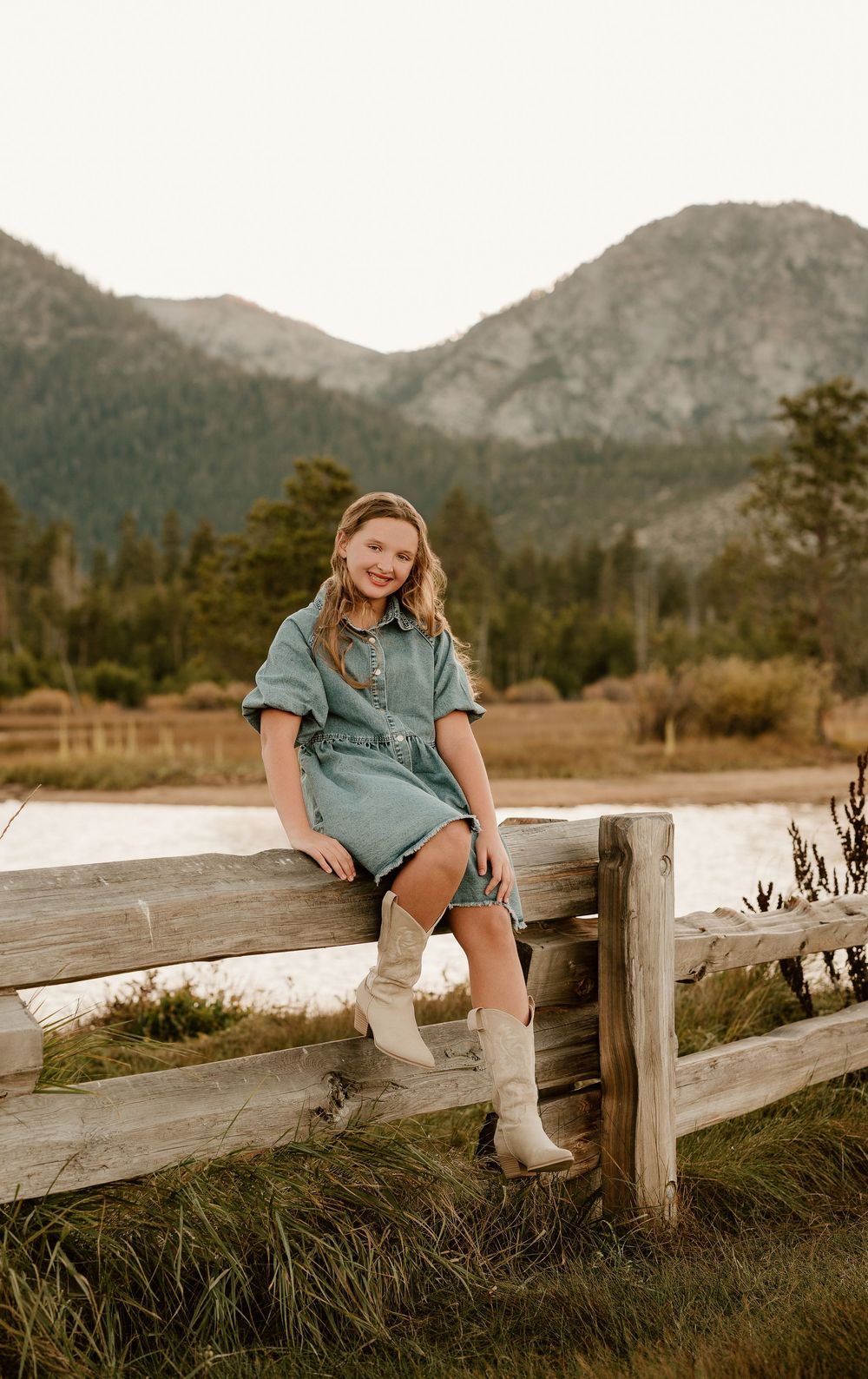 A person in denim sits on a wooden fence with mountains and meadow in the background.